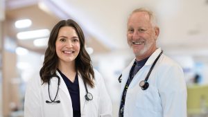 Two physicians in white coats stand side by side against a blurred clincal background